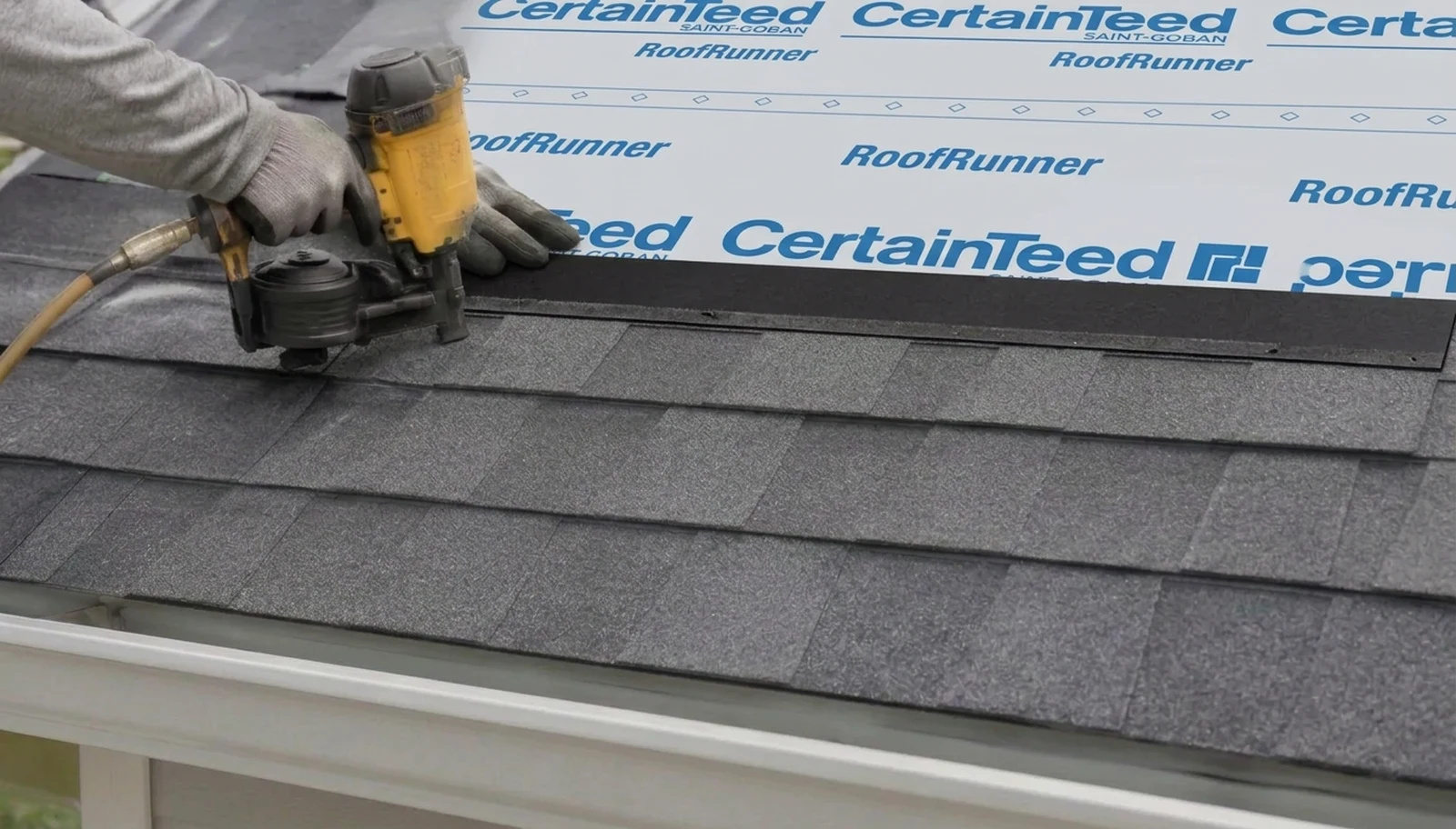 Gloved roofer fastening a shingle over synthetic underlayment, with staggered courses and a gutter below.